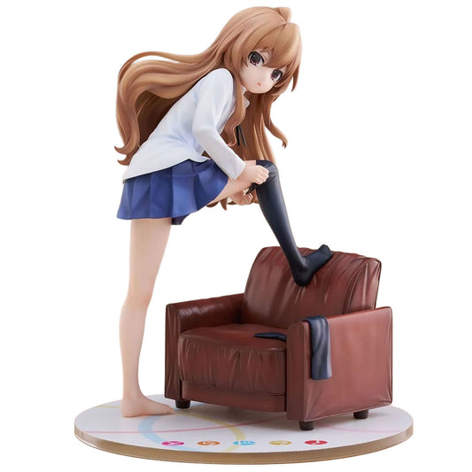An anime figurine of Taiga Aisaka from Toradora! with long light brown hair, wearing a white long-sleeve top, a blue pleated skirt, and black thigh-high socks, leaning over a small brown leather armchair and placing one foot inside the sock. The figure stands on a round display base against a plain white background.